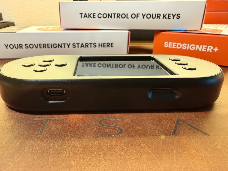 The bottom edge of the black SeedSigner+ Premium, with the USB-C power port on the left and the microSD slot on the right, photographed in front of stacked SeedSigner+ retail boxes